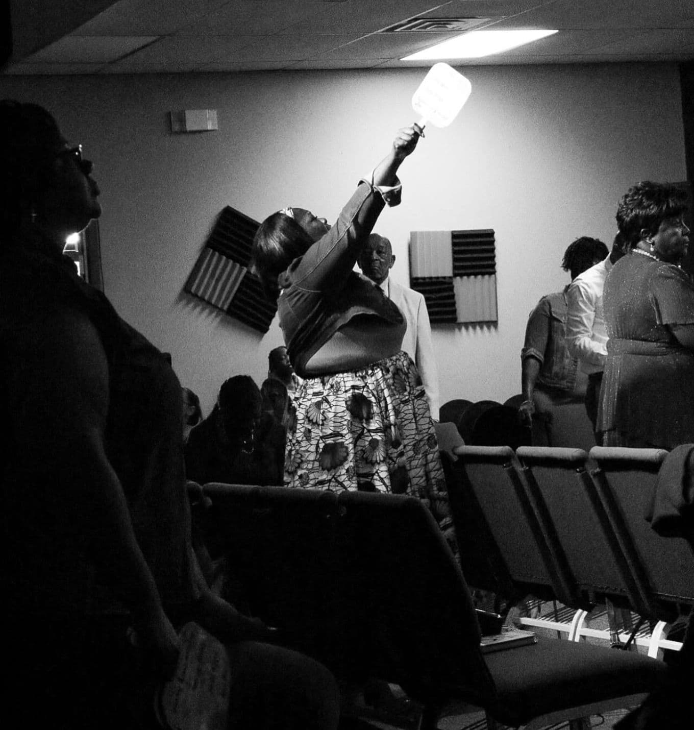 Black-and-white photo of worshippers standing among chairs at Advance Church, with a woman raising a hand holding a fan